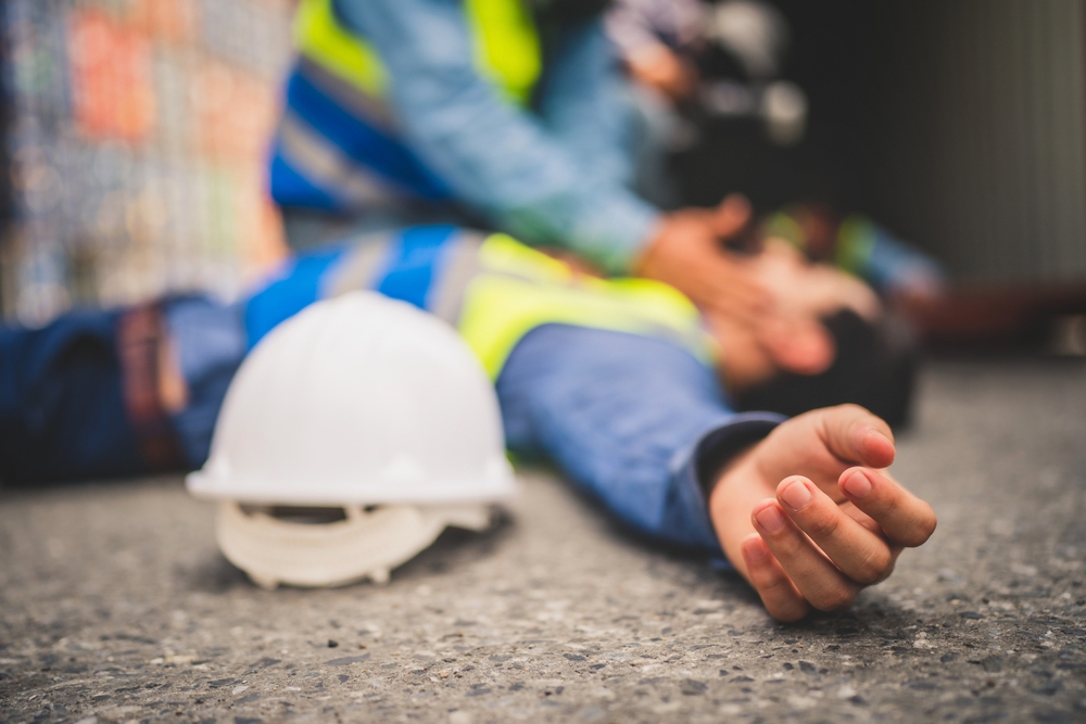 Injured construction worker lying on the ground while a coworker provides help beside a safety helmet.