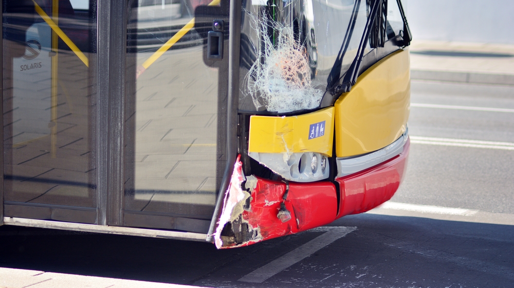 Front corner of a damaged public transit bus with a shattered windshield after a collision.